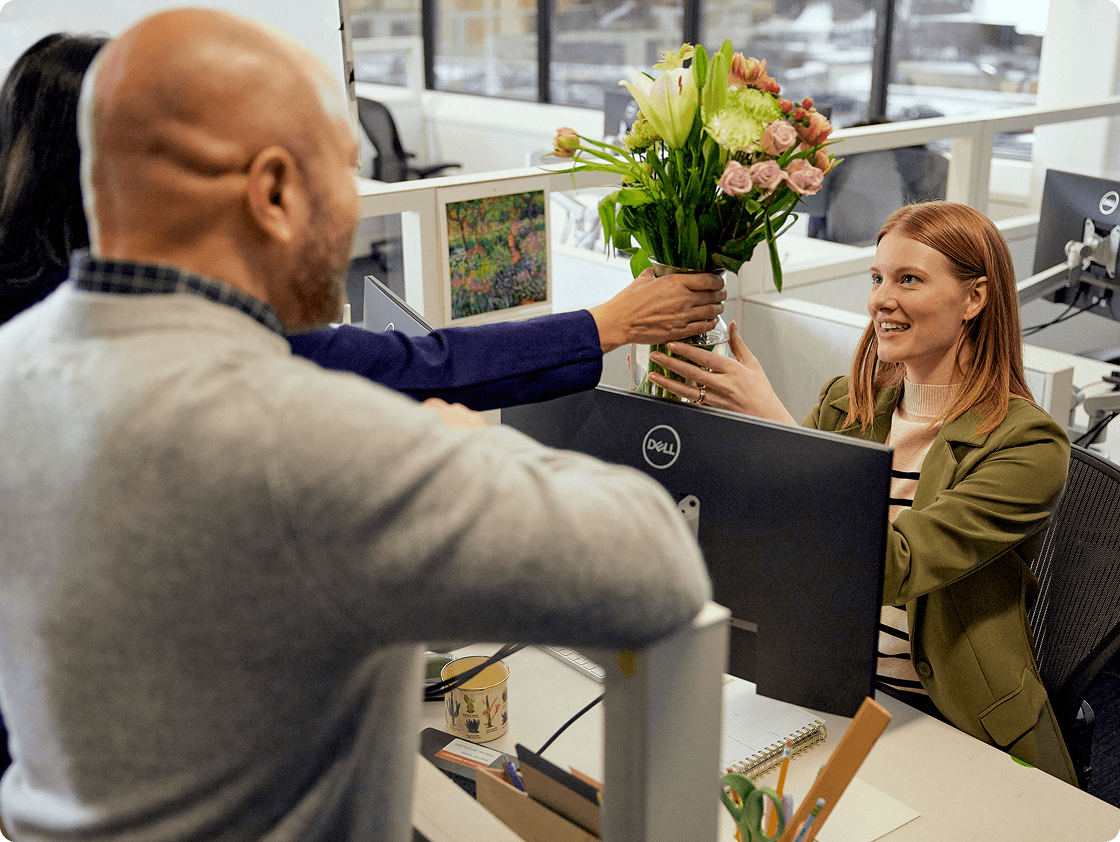 Image of Rose sitting at her desk at work and receiving flowers from a colleague on her first day back from leave. Devon is standing next to the colleage smiling.